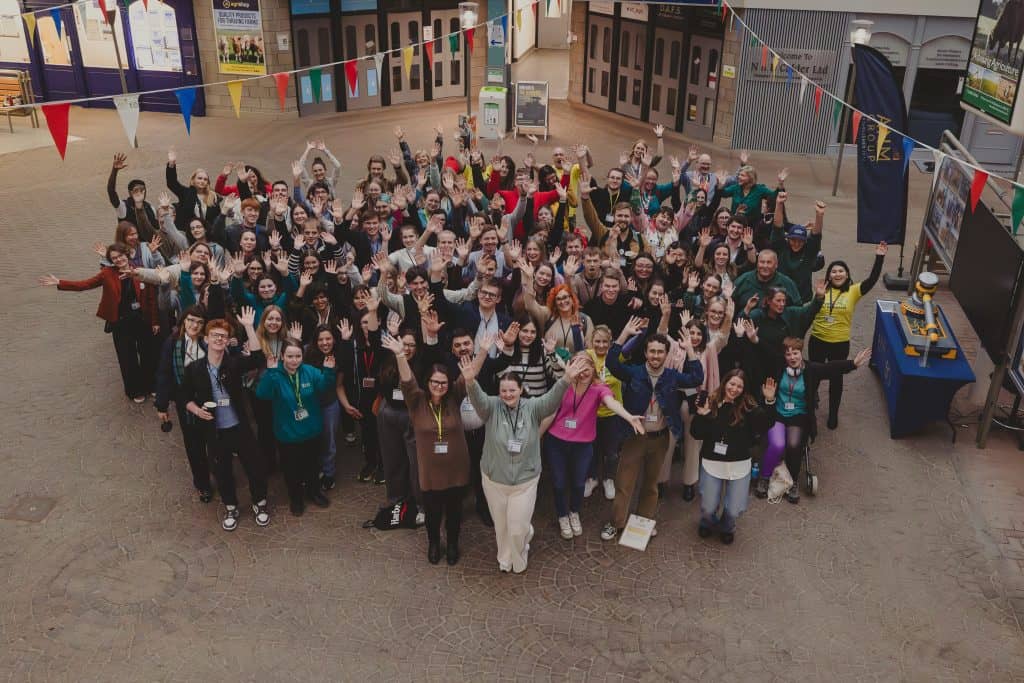 A large group of young people and community representatives gathered indoors, smiling and waving towards the camera at a Scottish Rural & Islands Youth Parliament event